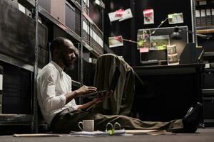 afrikansk amerikan polis Sammanträde på golv och arbetssätt på bärbar dator i kontor på natt tid. polis forskare granskning ledtrådar och misstänka foton på dator till lösa brottslighet fall