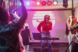 musiker sång och spelar elektronisk musik på skede i nattklubb. afrikansk amerikan kvinna dj använder sig av mikrofon och ljud kontrollant Utrustning medan utför i fullt med folk klubb foto