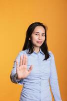 allvarlig filippinare kvinna höjning handflatan till annonsera sluta gest i studio över gul bakgrund, uttrycker avslag och vägran. modell som visar avslag gest, håller på med avslag tecken foto