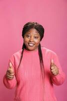 positiv afrikansk amerikan modell ger godkänd tecken stående över rosa bakgrund i studio. Lycklig glad kvinna med glad ansiktsbehandling uttryck som visar tummen upp gest, person håller på med tycka om tecken foto