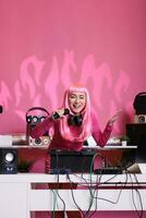 musiker stående på dj tabell innehav mikrofon talande med fläktar medan spelar techno låt använder sig av professionell mixer trösta. asiatisk artist med rosa hår njuter till prestera musik på natt i klubb foto