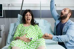 ung par förväntar sig bebis i sjukhus avdelning säng på sjukvård anläggningen. kvinna med graviditet har smärtsam sammandragningar få in i arbetskraft medan Make panik handla om förlossning. foto