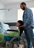 gravid kvinna Sammanträde i rullstol medan framtida far tar henne till moderskap rum framställning för barn leverans i sjukhus avdelning. ung par få redo för förlossning, diskuterar föräldraskap foto