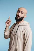 inspirerad arab man har aning och höjning index finger upp medan ser på kamera. ung skallig skäggig person med upphetsad ansiktsbehandling uttryck pekande uppåt studio porträtt foto