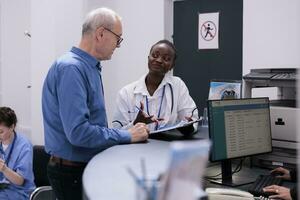 ung vuxen läkare sätta äldre patient till tecken medicinsk dokument medan diskuterar medicin behandling under kolla upp besök samråd i sjukhus väntar område. hälsa vård Stöd och begrepp foto