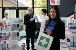leende asiatisk kvinna uppköp medicin i apotek och innehav inköp porträtt. apotek Lagra ung köpare stående nära hylla med läkemedel och bärande handla papper väska foto