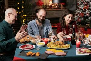 senior och ung människor på jul eve sammankomst på tabell till äta hemlagad mat och dryck vin, varelse festlig tillsammans. olika familj och vänner har roligt fira december Semester. foto