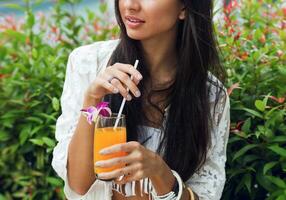 sensuell ung Lycklig kvinna avkopplande med gott färsk orange juice i trendig boho tropisk utrusta på henne semestrar. detaljer. friska livsstil. foto