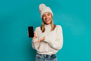 studio vinter- porträtt av leende blond kvinna i vit hatt och whool Tröja en innehav mobyle telefon , poserar i studio över turquose bakgrund. foto