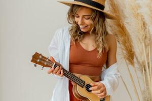 stänga upp Foto blond graciös kvinna i sommar utrusta med sugrör hatt spelar ukulele guitare över vit bakgrund i studio med pampas gräs dekor.