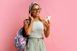 leende studerande kvinna i glasögon lyssnande musik förbi hörlurar och använder sig av mobyle telefon. stående på rosa bakgrund. bär eleganta sommar utrusta och tillbaka packa. foto