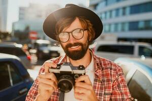 stänga upp porträtt av leende hipster skägg man använder sig av retro filma kamera, framställning foton medan reser. urban bakgrund. eleganta vår utrusta. bär svart hatt, rutig skjorta .