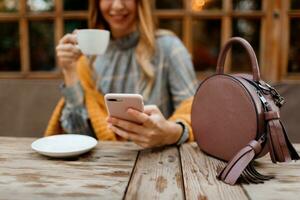 kvinna använder sig av mobil telefon, textning meddelande och dricka kaffe. eleganta väska på tabell. bär grå klänning och orange pläd. njuter mysigt morgon- i Kafé. foto