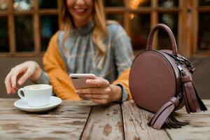 kvinna använder sig av mobil telefon, textning meddelande och dricka kaffe. eleganta väska på tabell. bär grå klänning och orange pläd. njuter mysigt morgon- i Kafé. foto