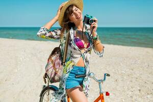 ung Lycklig kvinna i sommar badkläder och sugrör hatt med ryggsäck Framställ med årgång cykel på solig tropisk strand. innehav kamera. Söt turist njuter semester. foto