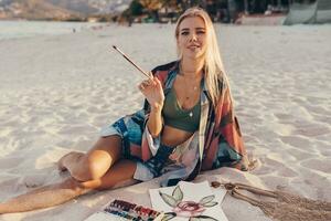 sommar livsstil bild av blond kvinna teckning vattenfärg blomma förbi borsta. konstnär Sammanträde på de strand. bohemisk utrusta. foto