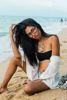 stänga upp livsstil porträtt av eleganta asiatisk kvinna med röd mun. hon är Sammanträde på solig tropisk strand. boho tillbehör.smycken ,armband och halsband. foto