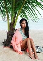 stänga upp sommar porträtt av ung skön asiatisk kvinna avkopplande på tropisk strand, bär boho trendig klänning, Sammanträde på vit sand nära handflatan träd. mode se. foto