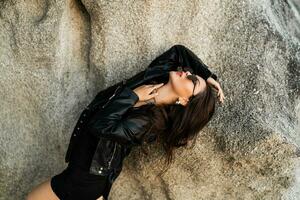 självsäker brunett kvinna i trendig svart läder jacka och sensuell svart klänning Framställ på klippig bakgrund. foto