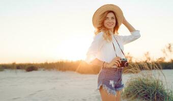 reser och högtider begrepp. Lycklig kvinna gående på kväll strand och innehav retro kamera. bär sugrör hatt och sexig jeans shorts. värma solnedgång färger . foto