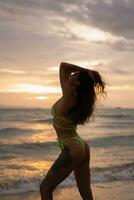 sexig solbränd kvinna stående och solbad på de strand. utomhus- Foto. sommar vibrafon. eleganta badkläder. solnedgång färger, vågig hårstrån. foto