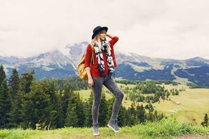 ung resande kvinna med hatt och ryggsäck njuter Fantastisk berg se ,gående och utforska verklig natur . blåsigt väder. bär eleganta boho falla utrusta, röd stickat Tröja och jeans. foto