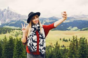 resande eleganta ung kvinna använda sig av smartphone tar själv Foto på bergen dal bakgrund, som visar tecken händer, grimaser, har roligt.