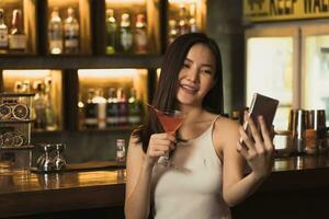 asiatisk kvinna som tar ett foto av sig själv medan hon dricker whisky i baren.