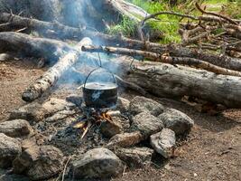 camping kök. matlagning mat på brand i vild. vattenkokare i sot hängande över brand. överlevnad i vild natur. underbar flamma med calderon. pott hänger i lägereld lågor. foto