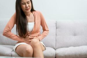 sjuk asiatisk kvinna lidande från akut abdominal smärta i de buk på grund av till menstruation period, pms. Sammanträde på soffa, magont från mat förgiftning, abdominal smärta, matsmältnings problem, gastrit foto