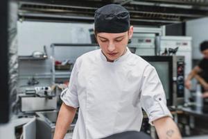 ung kock i vit uniform arbetar vid köket i restaurangen foto