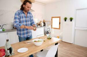 stilig man i tillfällig denim, stående på kök ö och framställning en friska rå vegan sallad i minimalistisk kök foto