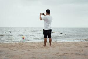 en avkopplande man stående på de strand och använder sig av en smartphone tar en Foto på de strand