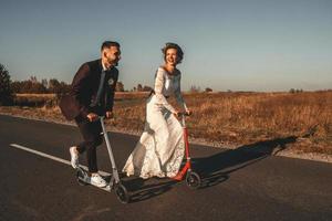 leende bröllopspar som åker på skoter längs vägen vid solnedgången foto