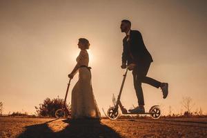 leende bröllopspar som åker på skoter längs vägen vid solnedgången foto