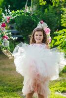 skön flicka i ett elegant rosa klänning hoppar nära gunga dekorerad med blommor. barn firar födelsedag 8 år. skön blommor vår tema gunga i trädgård foto