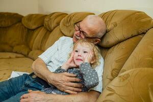 farfar kramar hans barnbarn Sammanträde på soffa. gammal man på Hem spelar med liten flicka. möte av ett äldre man och flicka efter isolering. foto