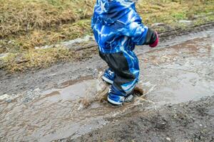 en barn promenader ner de bäck i de vår efter de snö smälter i de vår. en pojke i en blå overall och sudd stövlar i en pöl. foto