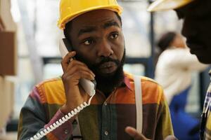 afrikansk amerikan man talande på fast telefon telefon i industriell magasin. fabrik lager operatör svar handledare telefon ring upp och lyssnande till lager förvaltning instruktioner foto