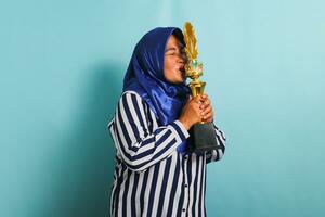 en Lycklig medelålders asiatisk affärskvinna i en blå hijab och en randig skjorta är kissing en guld trofé, fira henne Framgång och prestation. hon är isolerat på en blå bakgrund. foto