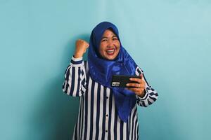 upphetsad medelålders asiatisk kvinna i en blå hijab och en randig skjorta, spelar en spel på mobil telefon, isolerat på en blå bakgrund. foto
