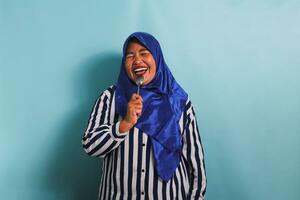 en Lycklig medelålders asiatisk kvinna i en blå hijab och en randig skjorta är innehav en sked och skrattande med glädje. hon är isolerat på en blå bakgrund foto
