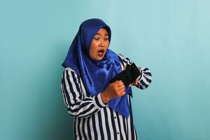 ett förvånad medelålders asiatisk kvinna i en blå hijab och en randig skjorta är ser in i henne tömma plånbok med en chockade och överraskad uttryck. hon är isolerat på en blå bakgrund foto