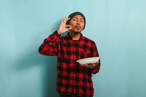 ung asiatisk man är framställning utsökt hand gest medan innehav tallrik, isolerat på blå bakgrund foto