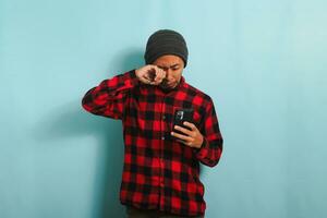 ett upprörd ung asiatisk man med en mössa hatt och en röd pläd flanell skjorta är gråt medan innehav en mobil telefon, upplever problem, och känsla arg och irriterad, isolerat på en blå bakgrund foto
