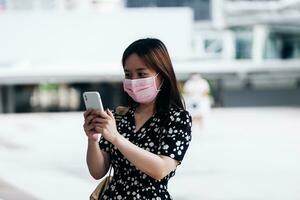 ung asiatisk kvinna bär ansikte mask använder sig av mobil telefon i de stad. foto
