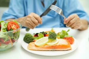 asiatisk äldre kvinna patient äter lax biff frukost med vegetabiliska friska mat medan Sammanträde och hungrig på säng i sjukhus. foto