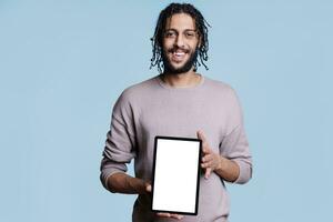leende arab man som visar digital läsplatta med vit tömma skärm för Ansökan annons falsk upp. Lycklig person presenter bärbar grej med tom pekskärm för produkt befordran foto