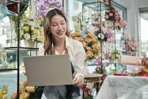 ett ung asiatisk kvinna blomsterhandlare och sme entreprenör i förkläde, Arbetar med bärbar dator, uppkopplad försäljning blommig ordna i färgrik blomma affär Lagra med skön knippa av blommar, och e-handel företag. foto