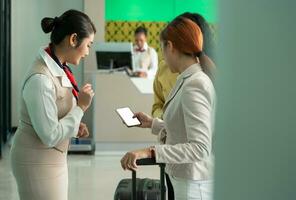 kvinna passagerare som visar flyg biljett till flygbolag personal på mobil telefon medan stående med bagage i flygplats terminal foto